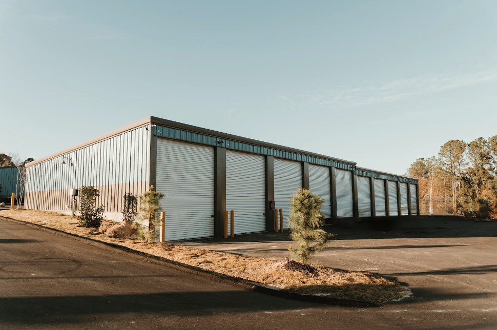 Three-quarter corner view of a large storage building with fresh landscaping and young pines under a clear blue sky