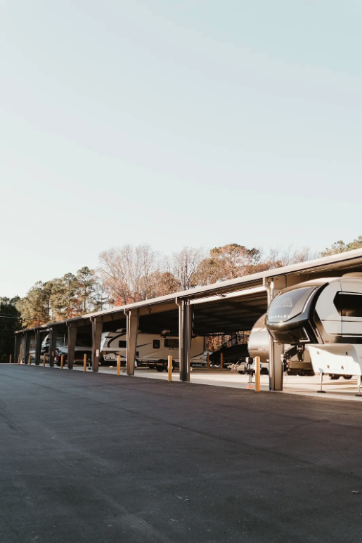 Long covered canopy parking bay at sunset with RVs and a large fifth-wheel trailer parked underneath on a wet asphalt drive
