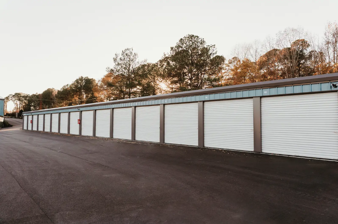 Long row of drive-up units with white roll-up doors and blue-green trim on a wet asphalt drive at sunset