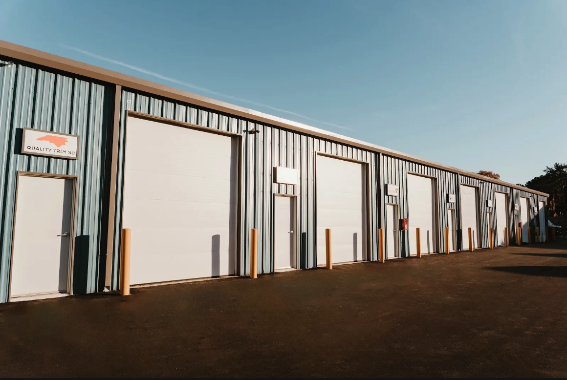 Row of commercial flex and warehouse units with large white overhead doors, man-doors, and yellow bollards under a clear blue sky