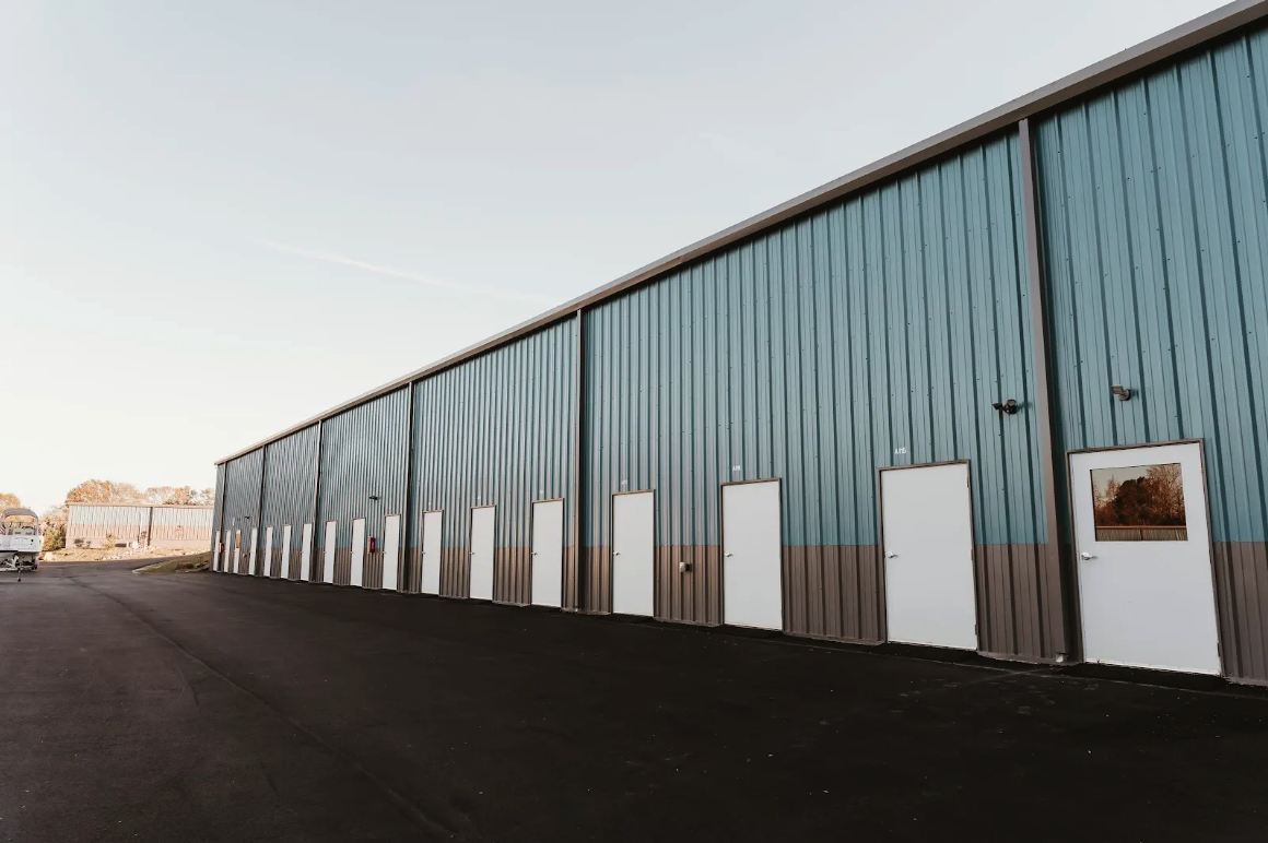 Large teal metal warehouse-style building with white man-doors along a clean asphalt drive at dusk
