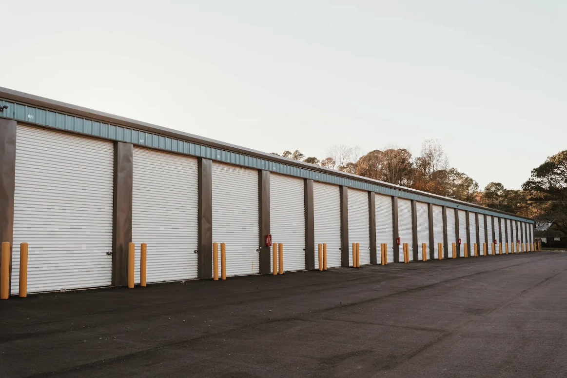 Long facade of white roll-up doors lined with yellow bollards along a wide asphalt aisle at dusk