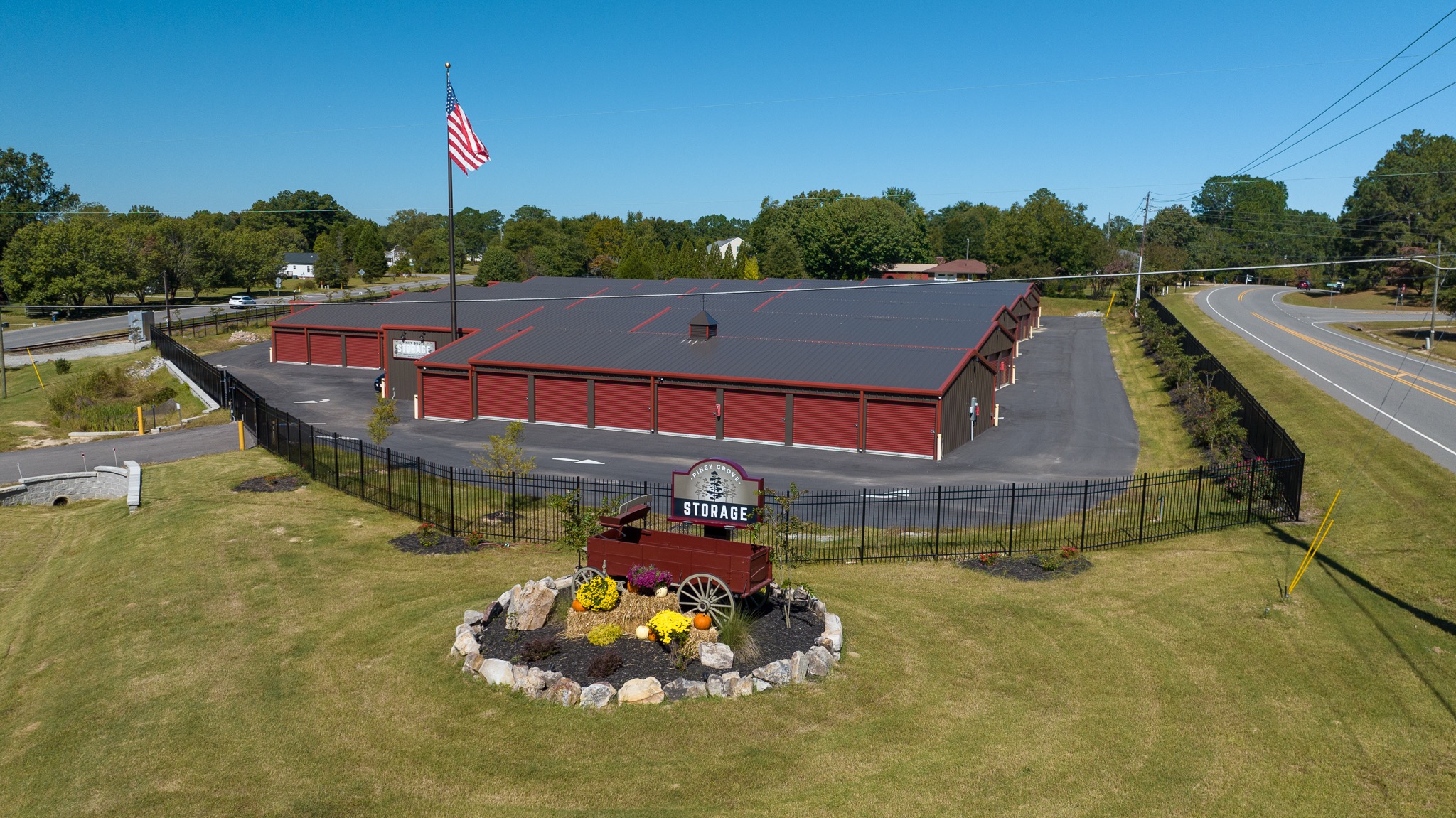 Aerial view of the full Piney Grove Storage facility with red drive-up buildings inside a black security fence, the monument sign and flag out front, under a clear blue sky