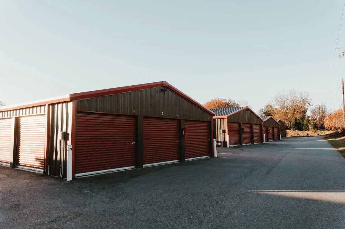 A row of drive-up storage buildings with maroon roll-up doors and brown metal siding under gabled roofs along a wide asphalt drive at golden hour